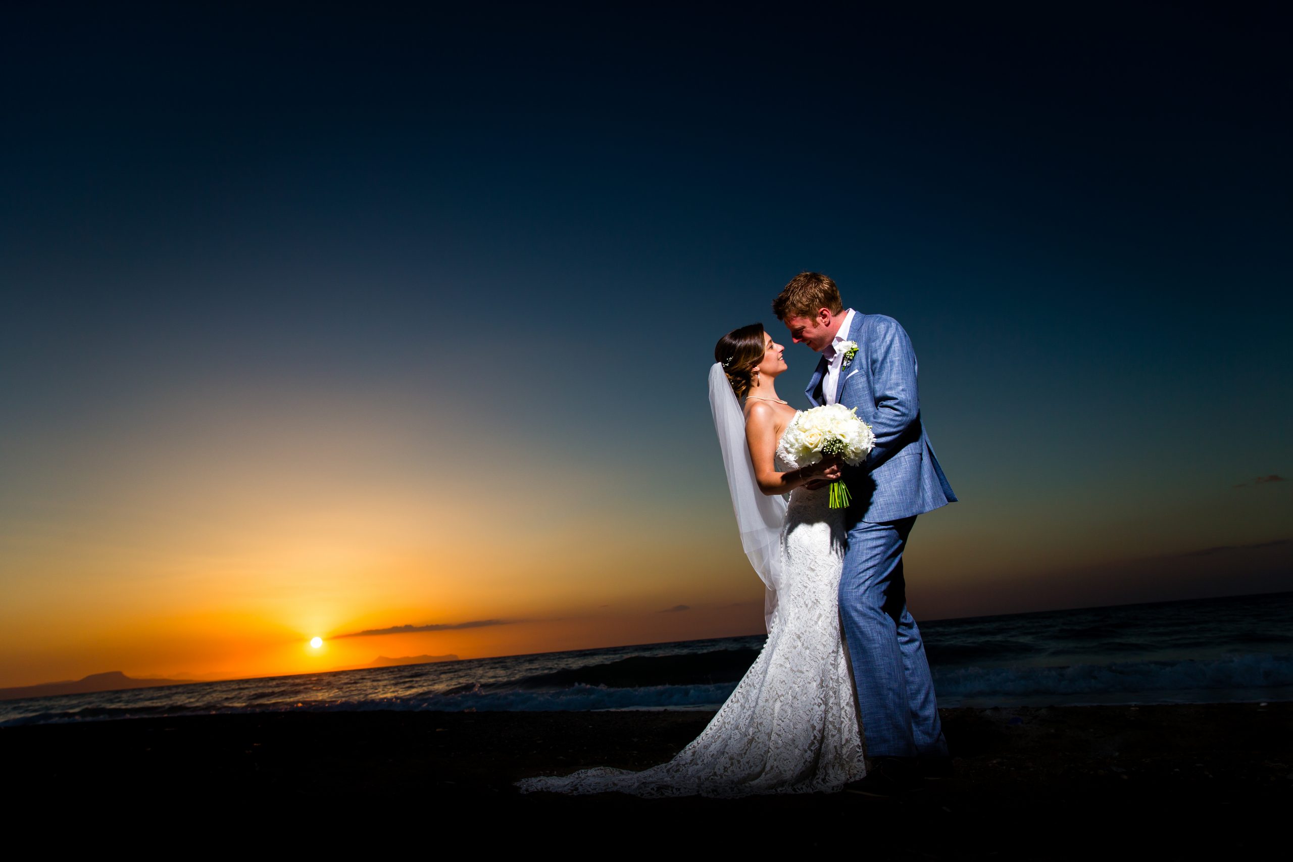 Fashion-style portrait of a wedding couple in Crete at sunset, elegantly posed with warm golden light, soft sea views, and a romantic, editorial feel.